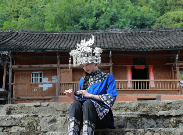 图片2身着水族传统服饰的潘安琪同学，背景为家乡吊脚楼.png